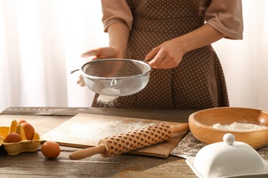 Photo of Woman sifting flour at wooden table with rolling pin indoors, closeup