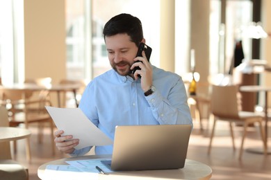 Photo of Handsome business owner talking by smartphone at table with laptop in his cafe