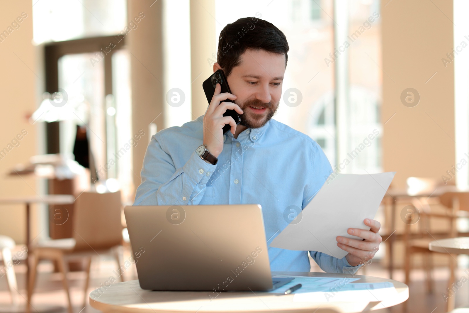 Photo of Handsome business owner talking by smartphone at table with laptop in his cafe