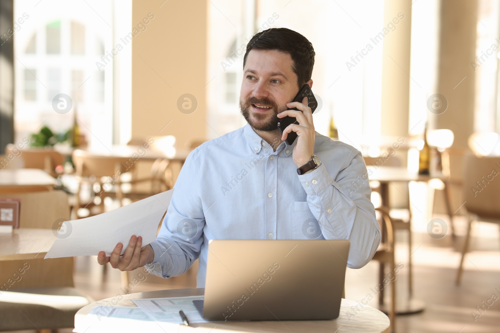 Photo of Smiling business owner talking by smartphone at table with laptop in his cafe