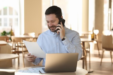 Smiling business owner talking by smartphone at table with laptop in his cafe