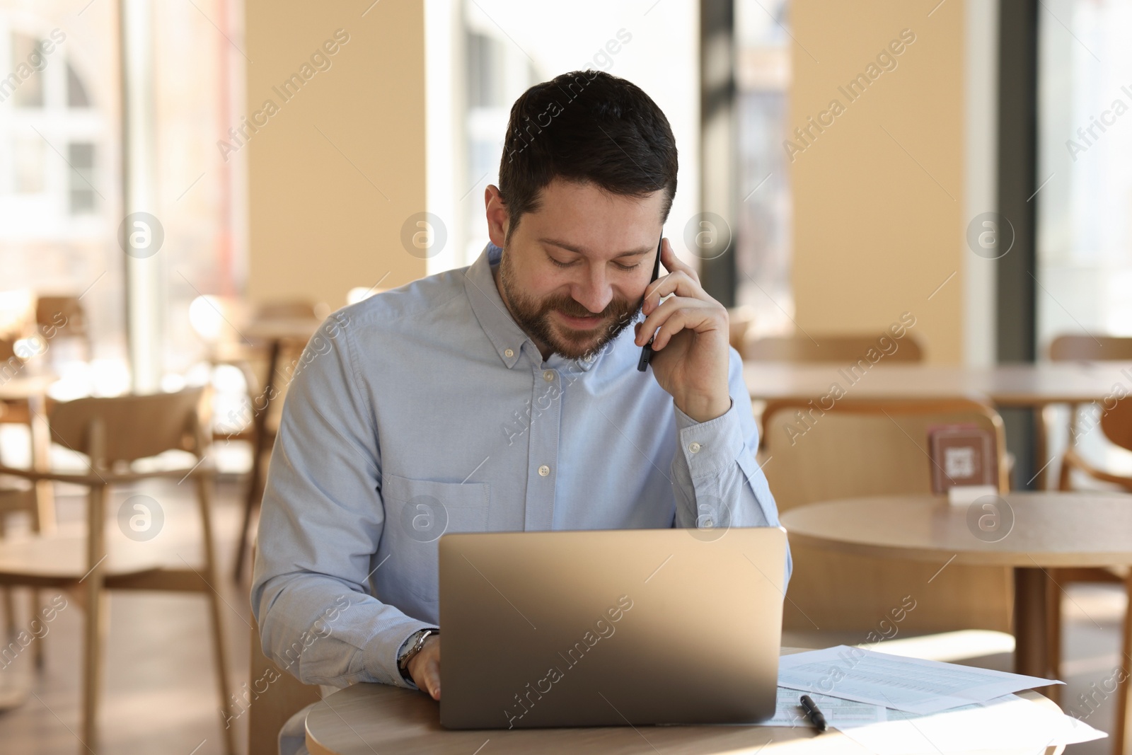 Photo of Handsome business owner talking by smartphone at table with laptop in his cafe