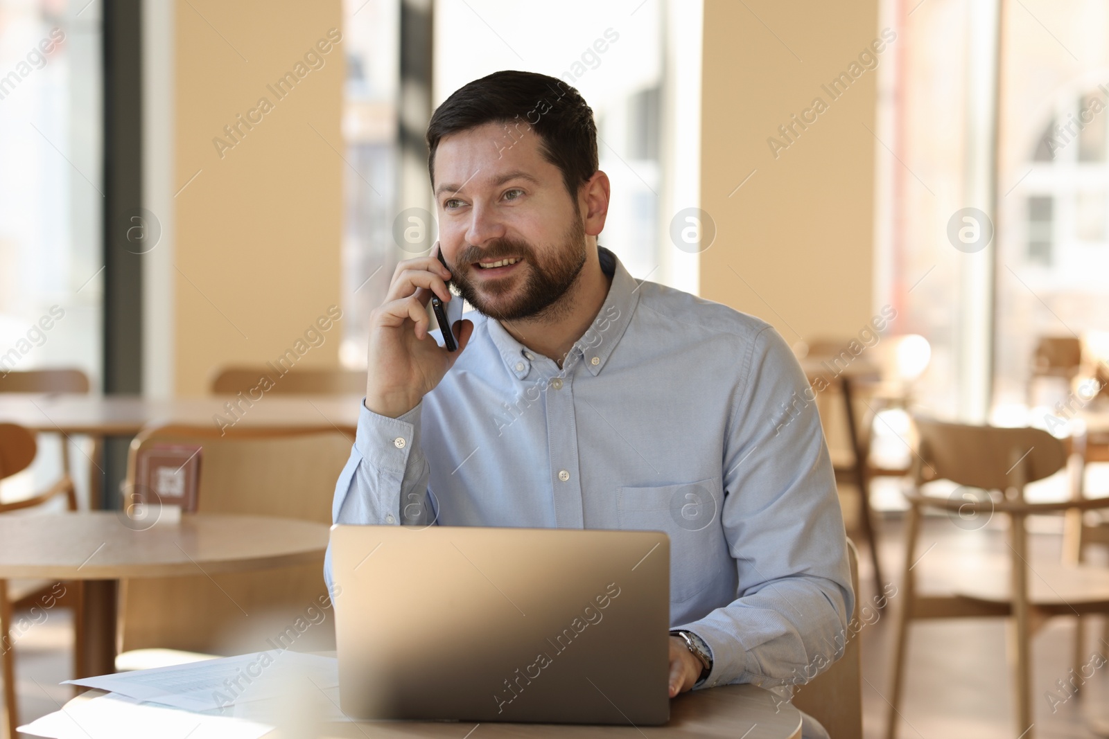 Photo of Smiling business owner talking by smartphone at table with laptop in his cafe