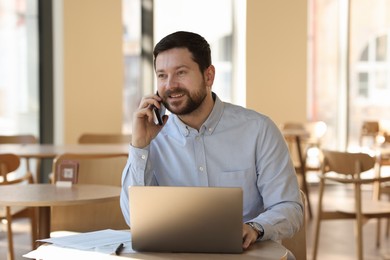 Photo of Smiling business owner talking by smartphone at table with laptop in his cafe