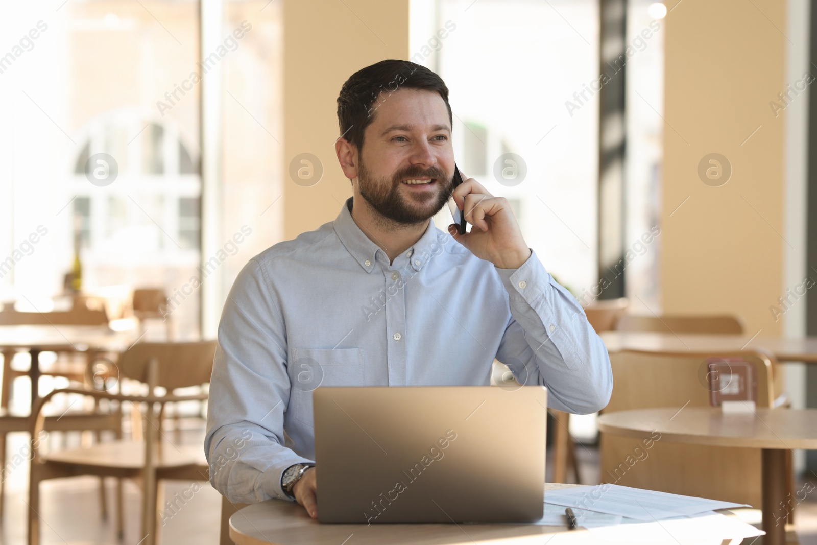 Photo of Smiling business owner talking by smartphone at table with laptop in his cafe