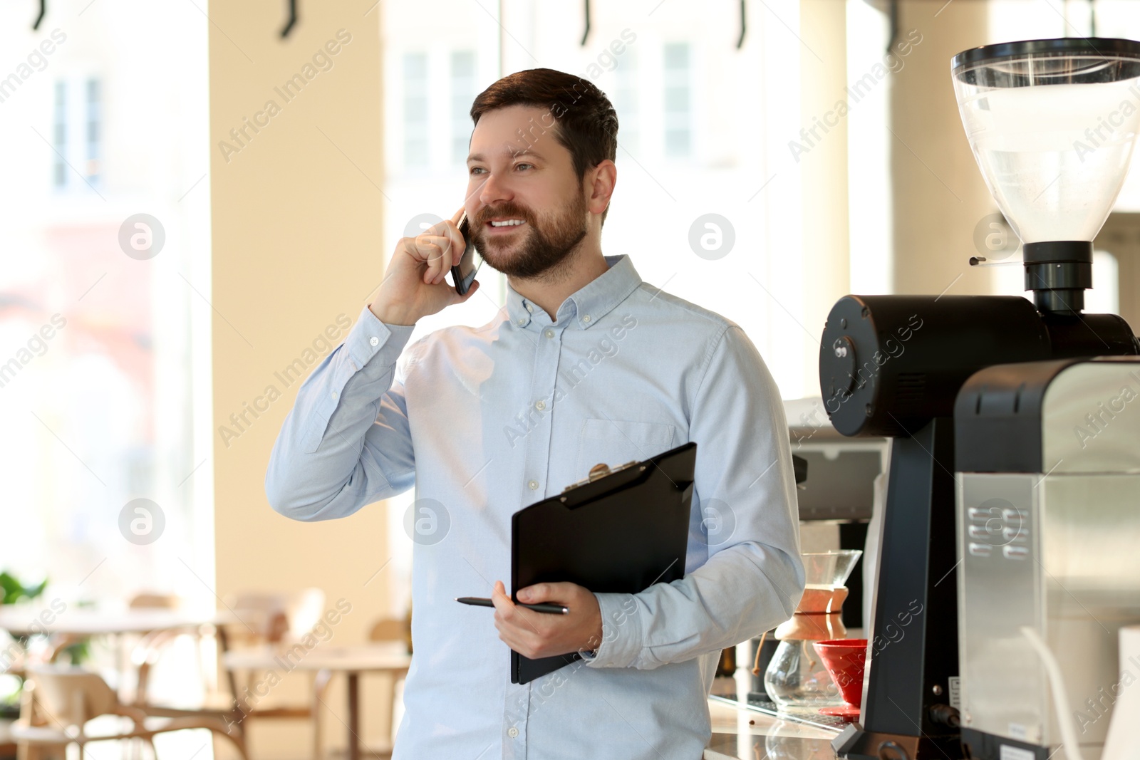 Photo of Smiling business owner with clipboard talking by smartphone in his cafe