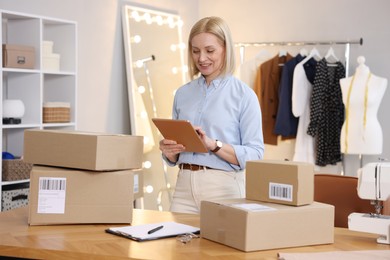 Photo of Business owner using tablet near table with parcels in her tailor shop