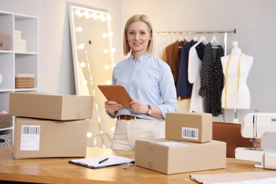 Photo of Business owner using tablet near table with parcels in her tailor shop