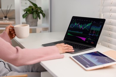 Photo of Stock exchange. Woman with cup of drink analysing financial market on laptop at white table indoors, closeup