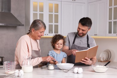 Photo of Cute little girl with her father and grandmother cooking by recipe book in kitchen