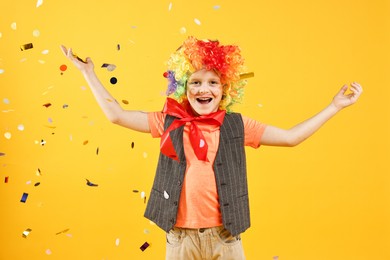 Happy little boy dressed like clown and flying confetti on orange background. Surprise party