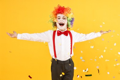 Happy little boy dressed like clown and flying confetti on yellow background. Surprise party