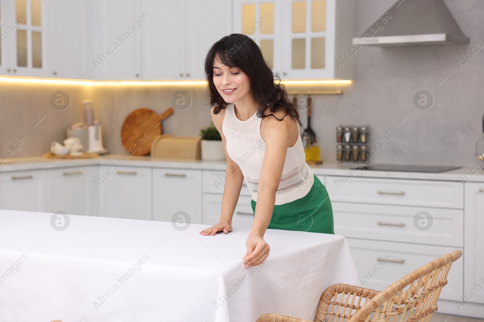 Photo of Young woman putting white tablecloth on table in kitchen