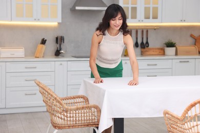 Young woman putting white tablecloth on table in kitchen