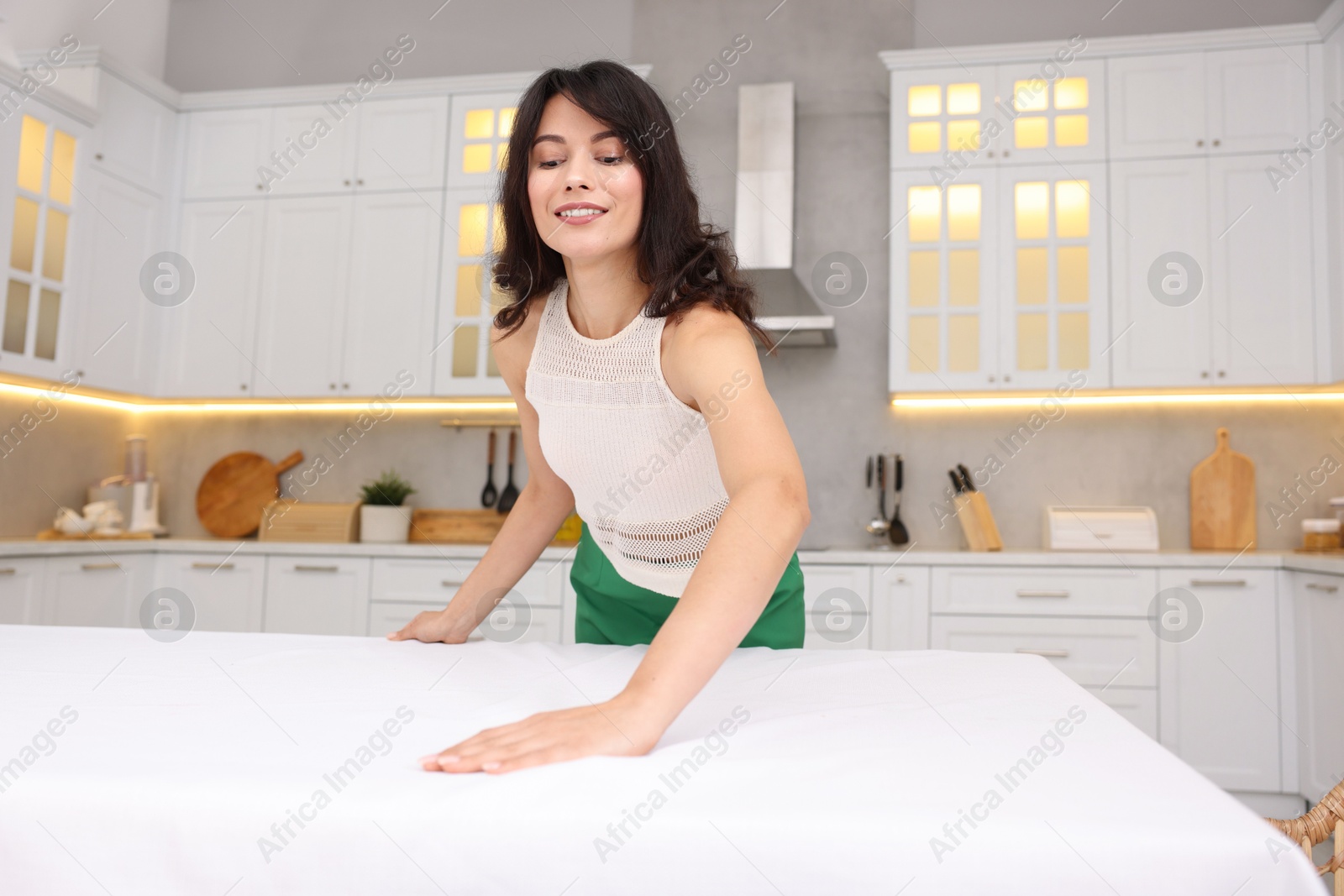 Photo of Young woman putting white tablecloth on table in kitchen, low angle view