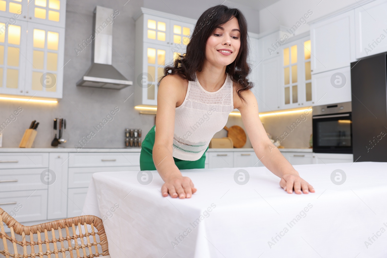 Photo of Young woman putting white tablecloth on table in kitchen, low angle view