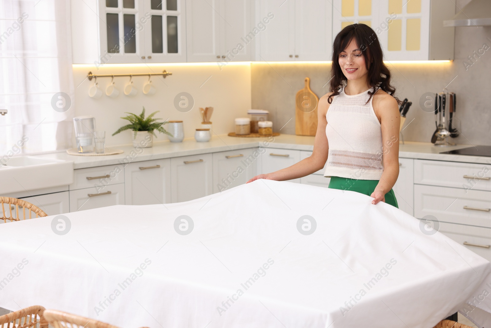 Photo of Young woman putting white tablecloth on table in kitchen