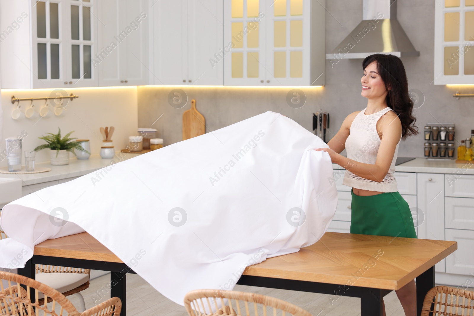 Photo of Young woman putting white tablecloth on table in kitchen