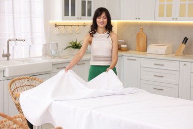 Young woman putting white tablecloth on table in kitchen