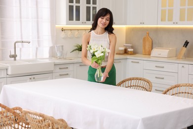 Photo of Woman putting vase with flowers on table with white tablecloth in kitchen