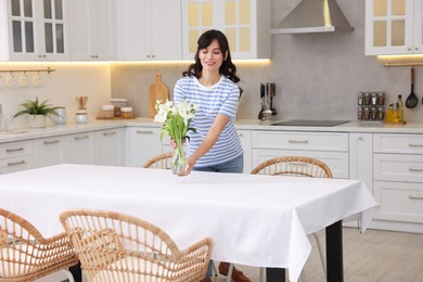 Photo of Woman putting vase with flowers on table with white tablecloth in kitchen