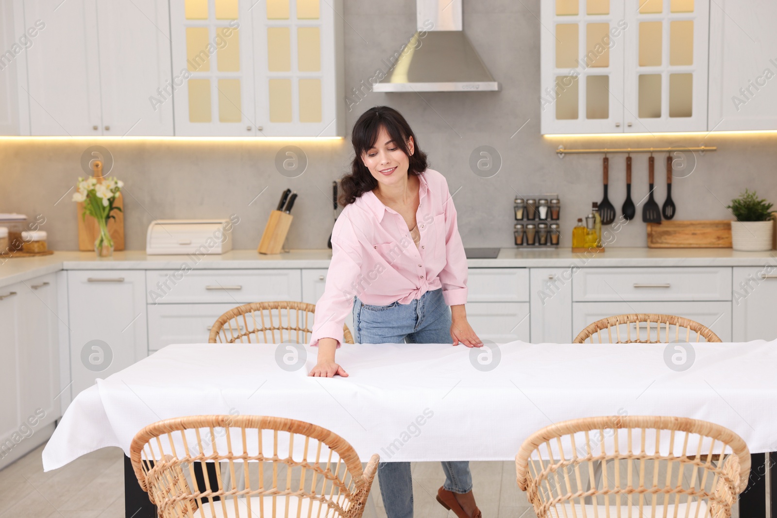 Photo of Young woman putting white tablecloth on table in kitchen