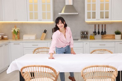 Young woman putting white tablecloth on table in kitchen