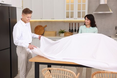 Couple putting white tablecloth on table in kitchen