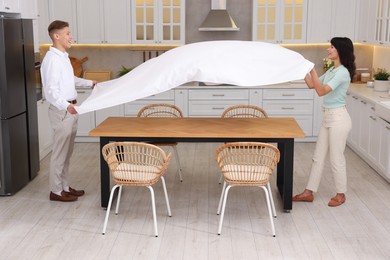 Couple putting white tablecloth on table in kitchen