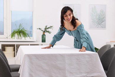 Young woman putting white tablecloth on table at home