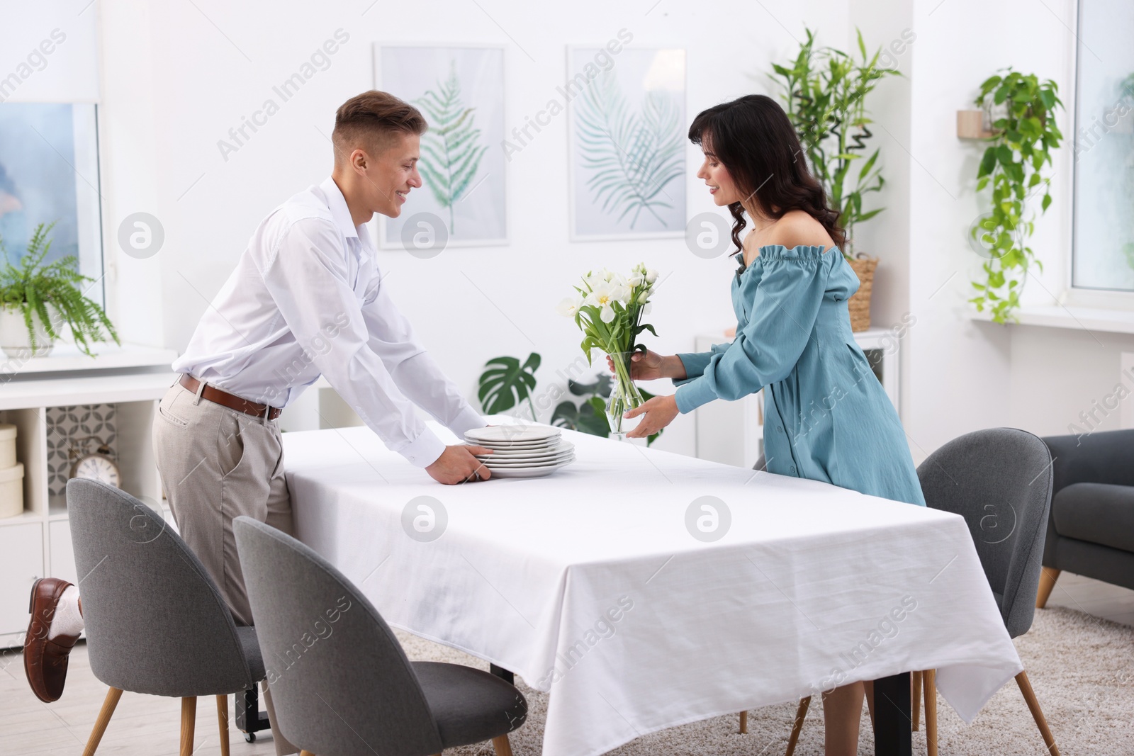 Photo of Couple putting plates and vase with flowers on table with tablecloth at home