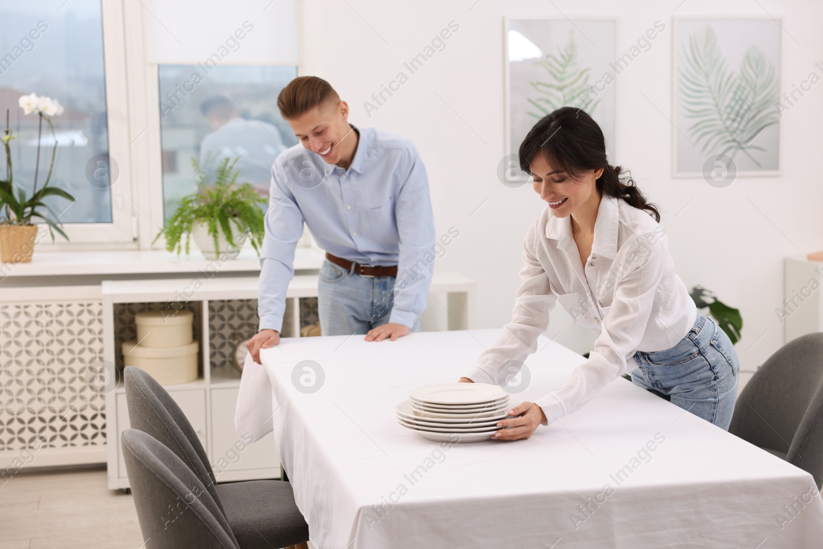 Photo of Couple putting plates on table with white tablecloth at home