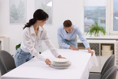 Couple putting plates on table with white tablecloth at home, selective focus