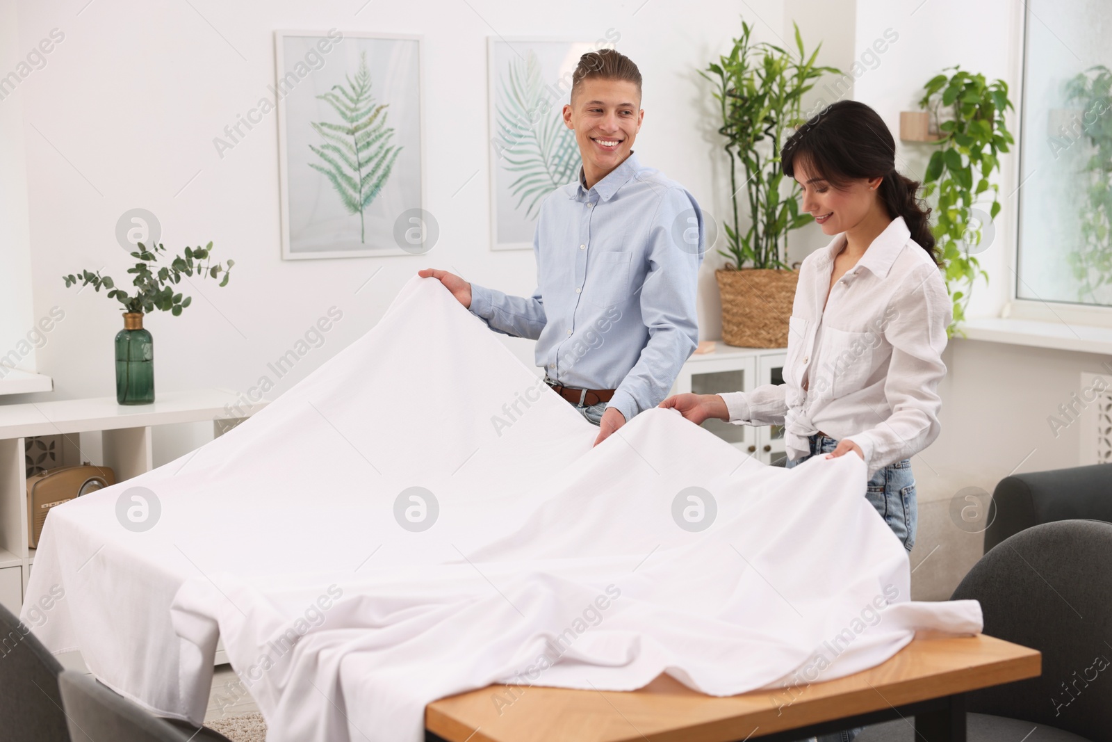 Photo of Couple putting white tablecloth on table at home