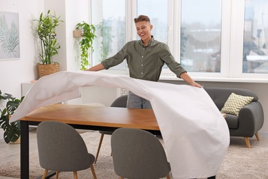 Young man putting white tablecloth on table at home