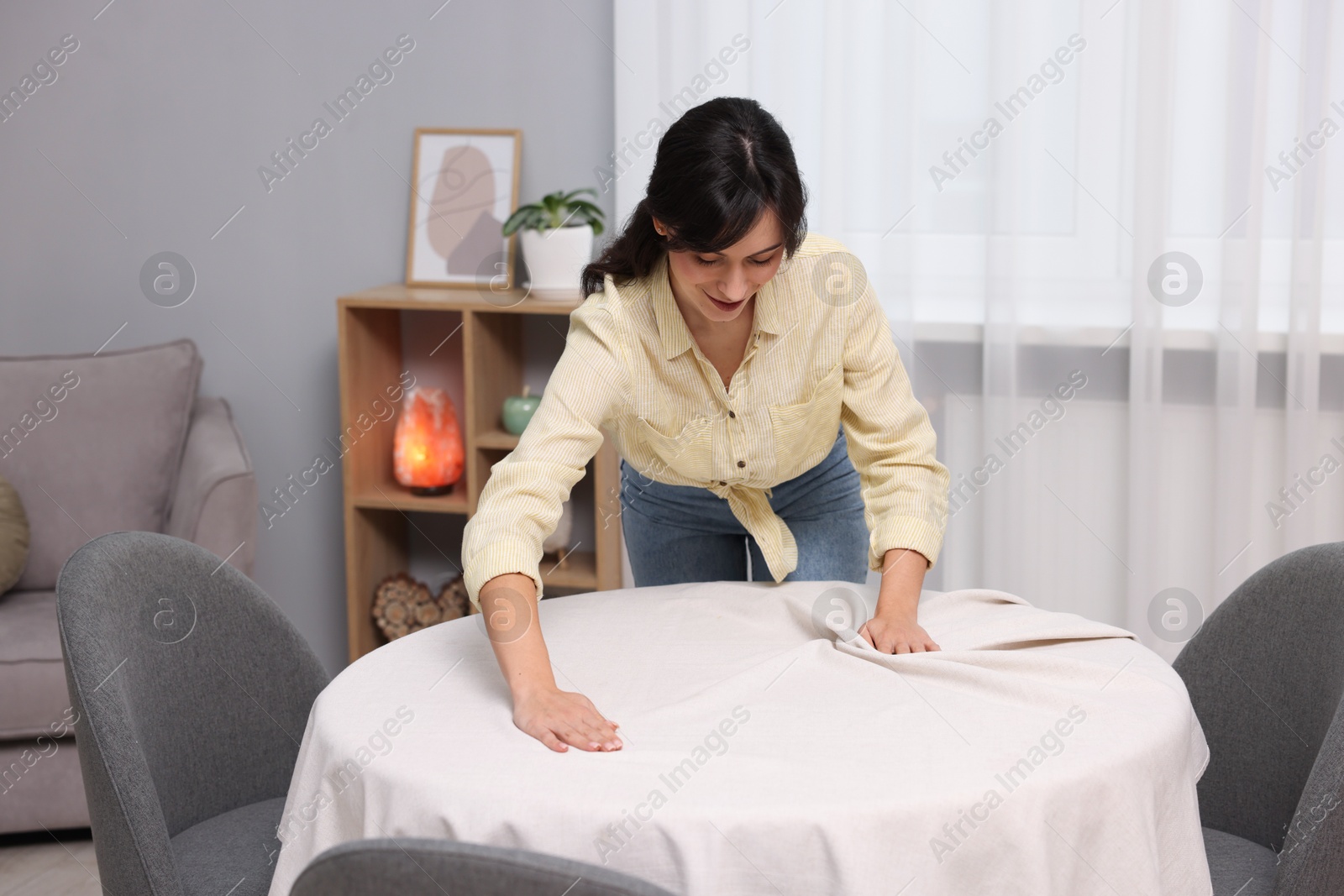 Photo of Young woman putting white tablecloth on table at home