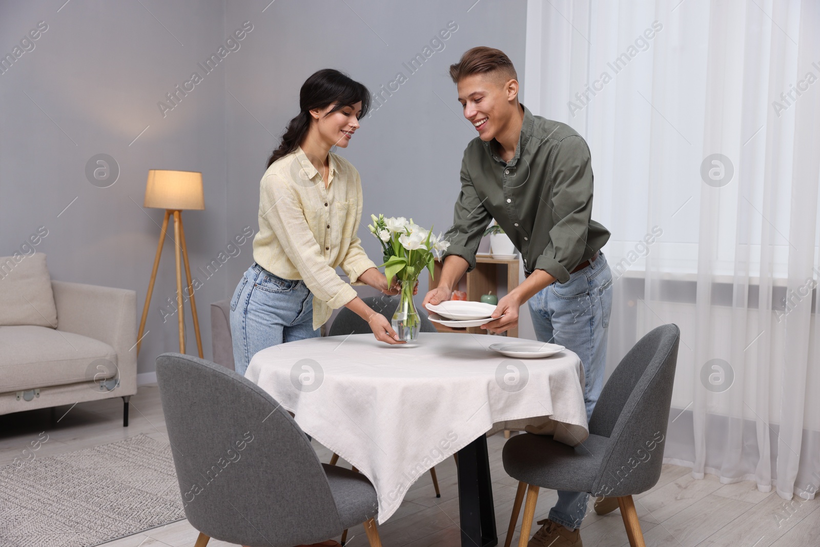 Photo of Couple putting plates and vase with flowers on table with white tablecloth at home