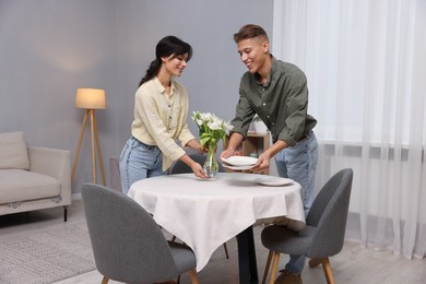 Photo of Couple putting plates and vase with flowers on table with white tablecloth at home