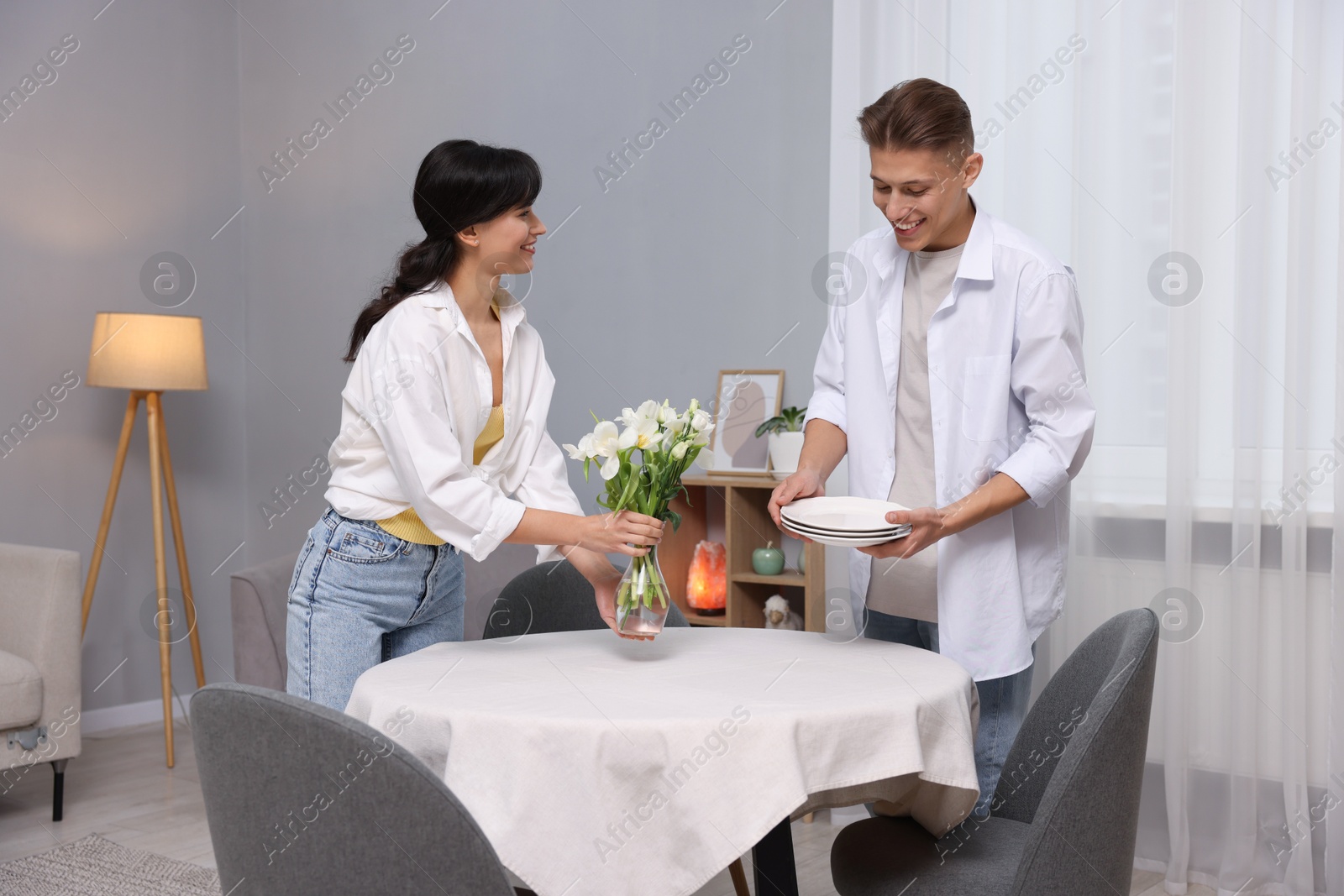 Photo of Couple putting plates and vase with flowers on table with white tablecloth at home