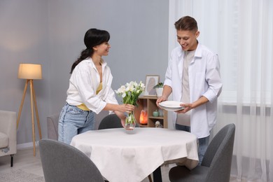 Couple putting plates and vase with flowers on table with white tablecloth at home