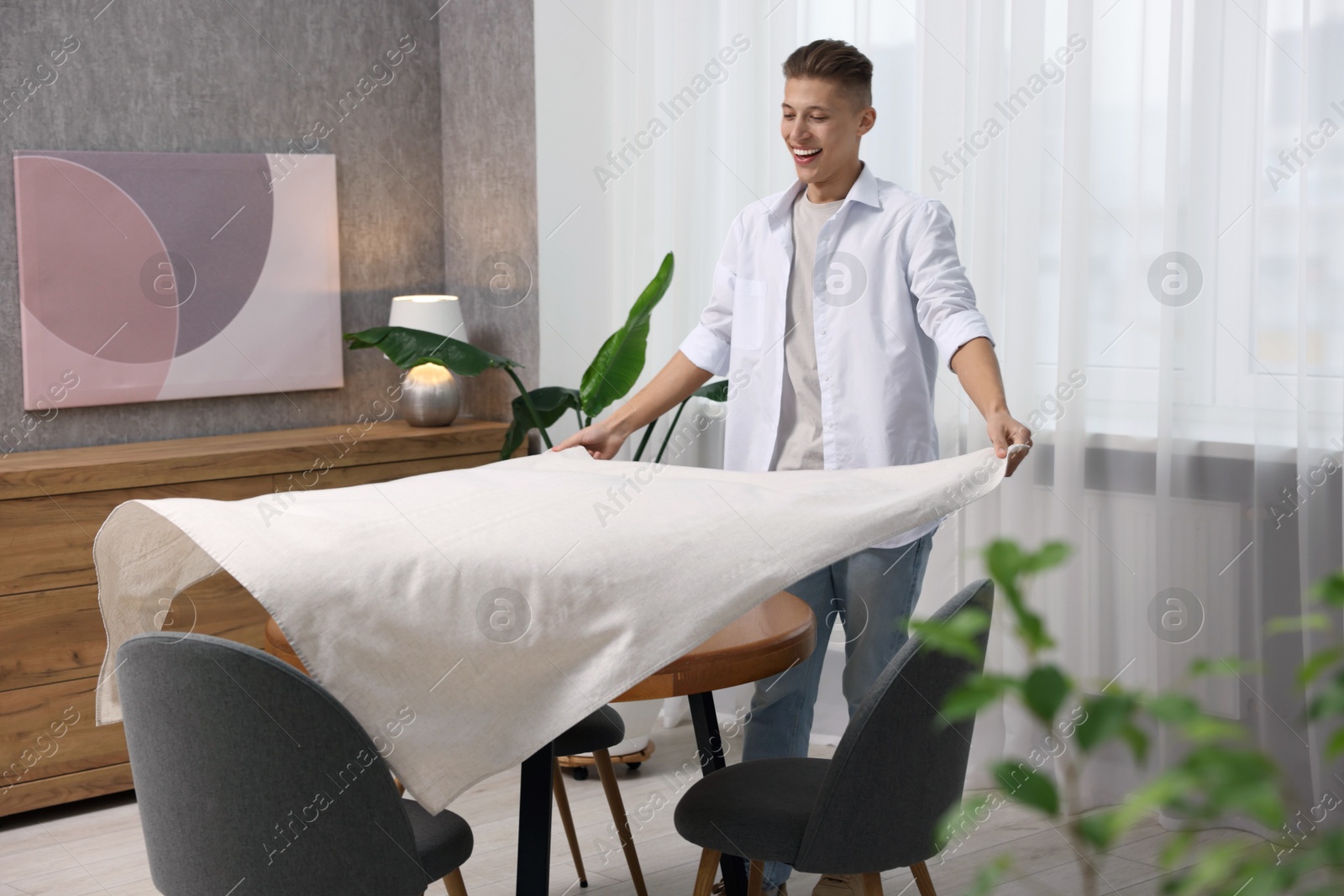 Photo of Young man putting white tablecloth on table at home