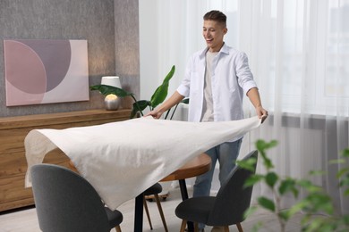 Young man putting white tablecloth on table at home