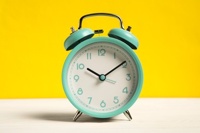 Photo of One turquoise alarm clock on white wooden table against yellow background