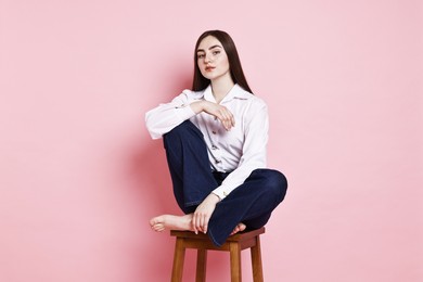 Photo of Beautiful young woman in stylish jeans sitting on stool against pink background