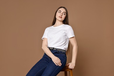 Photo of Beautiful young woman in stylish jeans sitting on stool against brown background