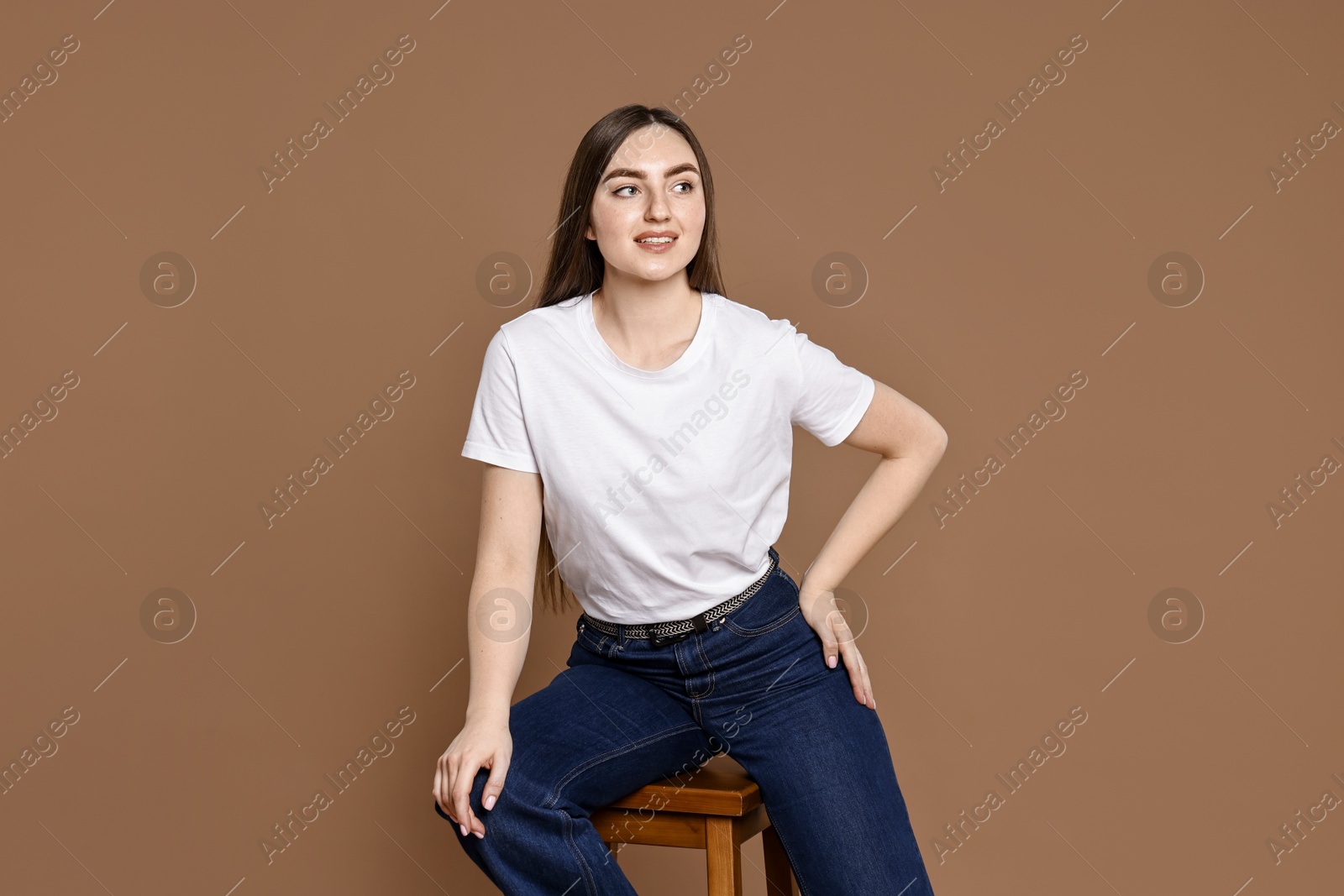 Photo of Smiling woman in stylish jeans sitting on stool against brown background