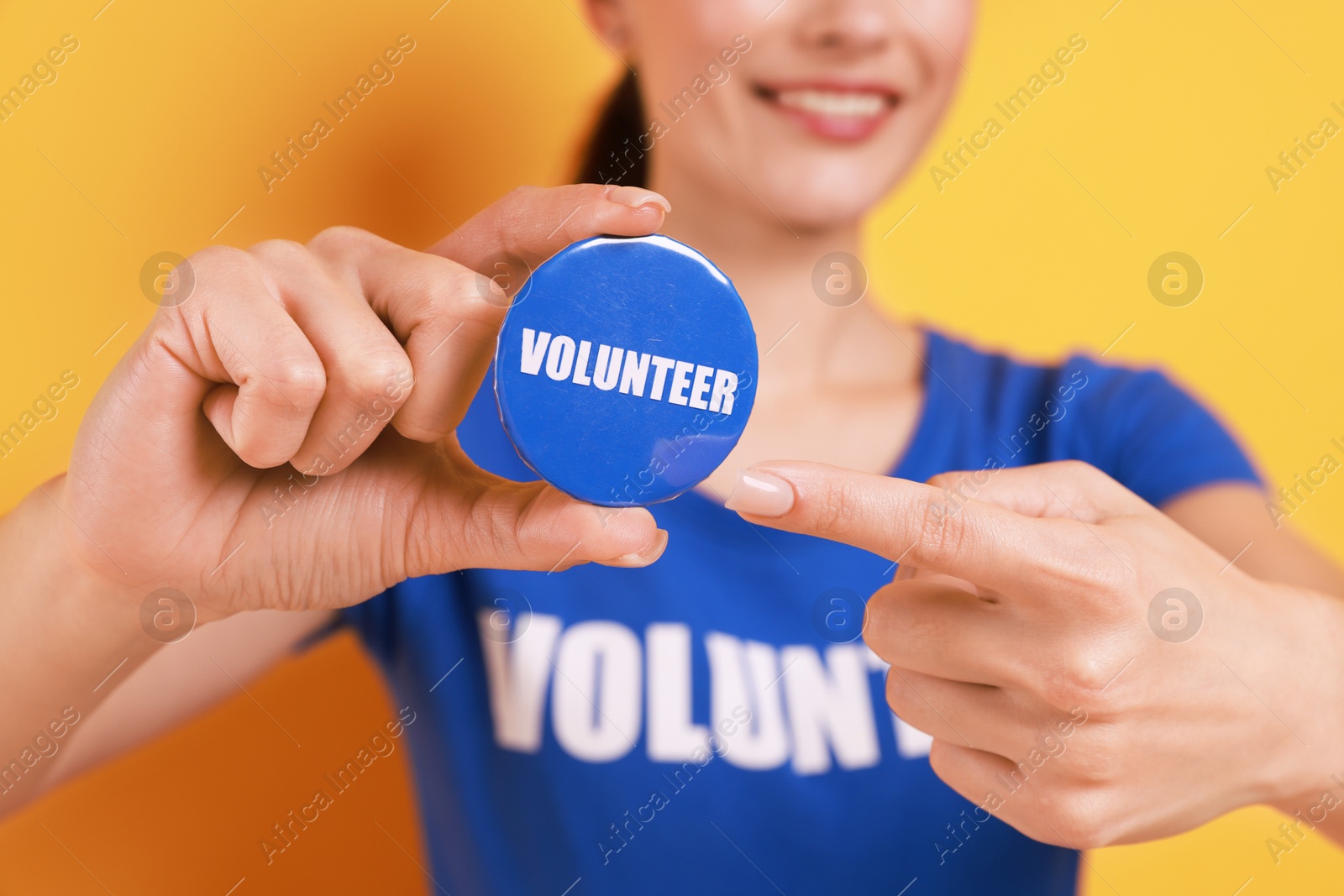 Photo of Woman pointing at blue button badge with word Volunteer on orange background, closeup