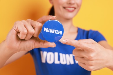 Woman pointing at blue button badge with word Volunteer on orange background, closeup