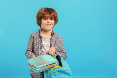 Little boy packing lunch box into backpack on light blue background. Space for text