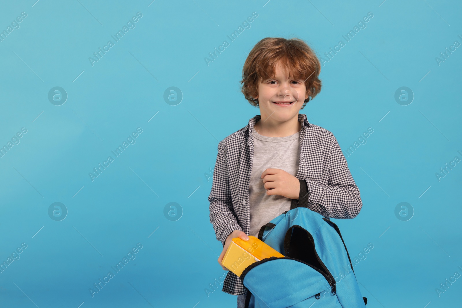 Photo of Little boy packing lunch box into backpack on light blue background. Space for text